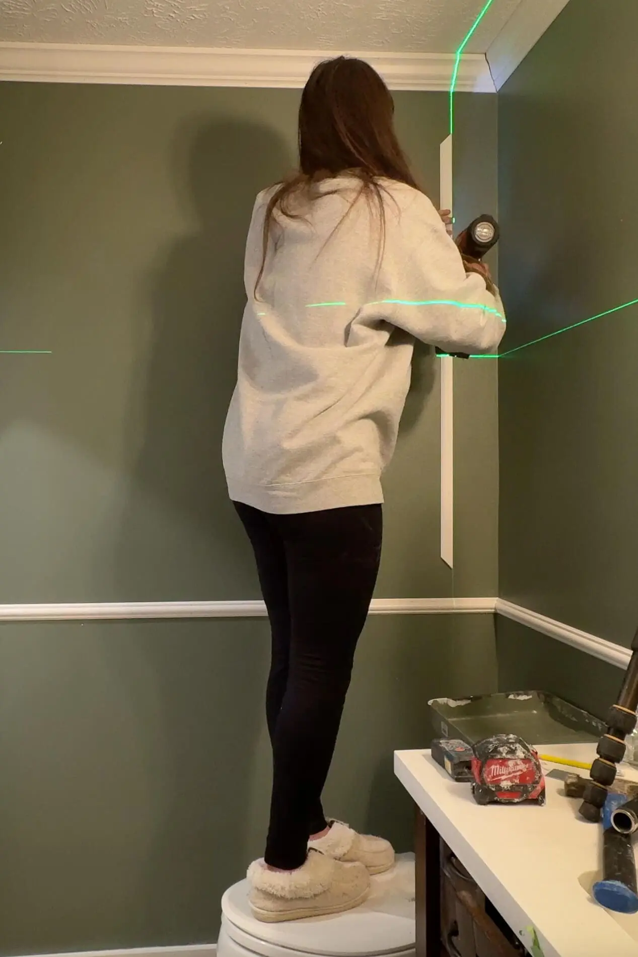 Person standing on a stool installing vertical trim pieces on a green bathroom wall using a nail gun, creating DIY picture frame molding with precise alignment.